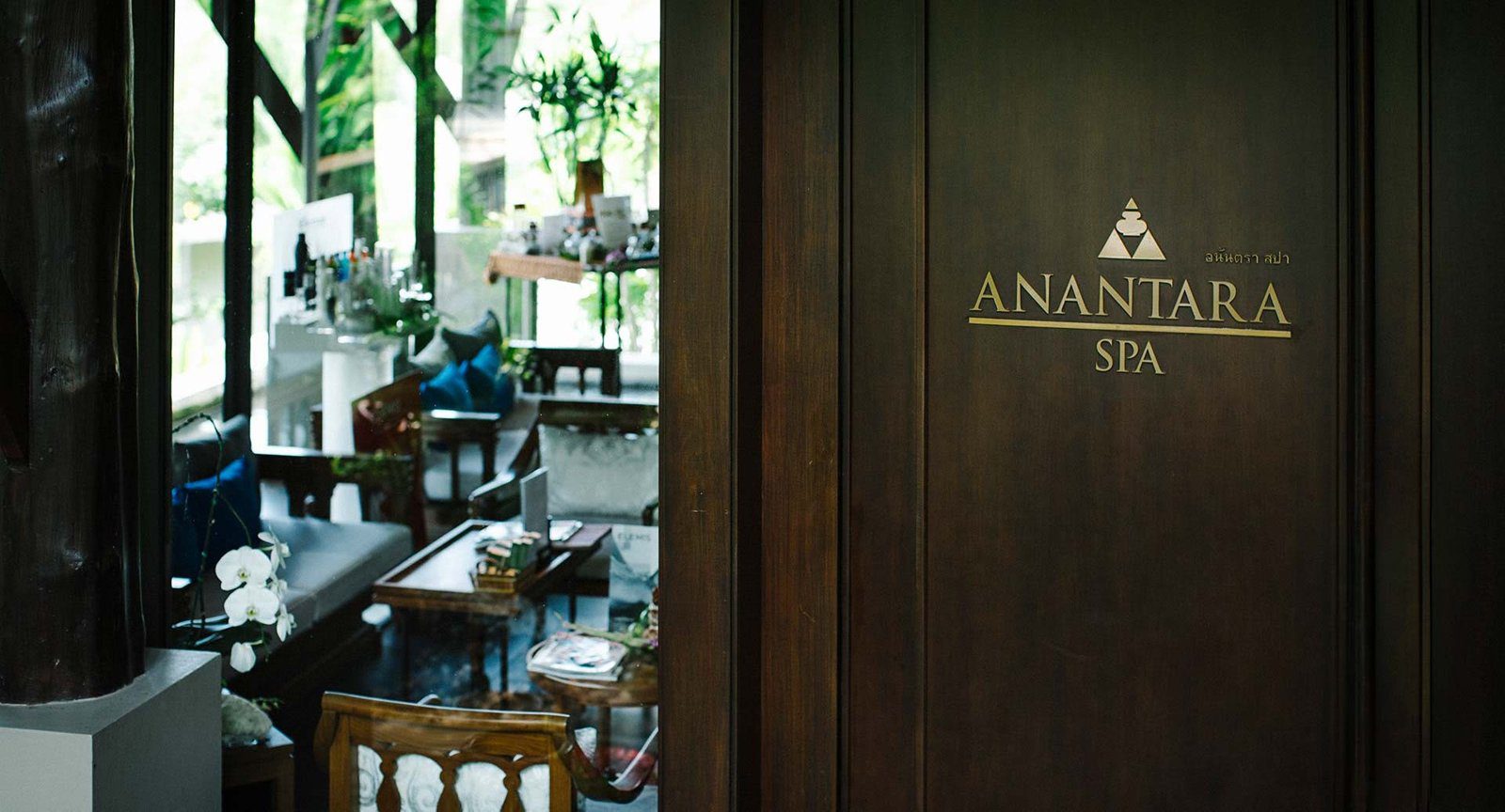 深色木門上寫著 ANANTARA SPA 金色字樣，門內是擺著沙發與蘭花的接待區
