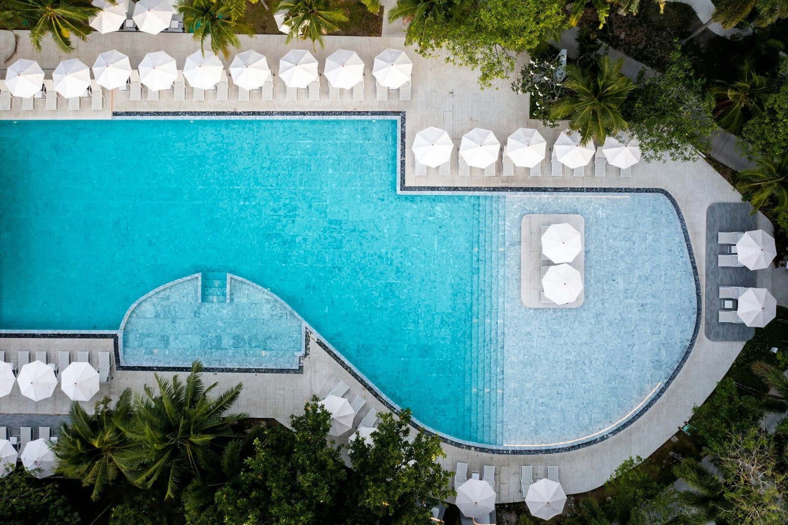空拍 Tropicale Pool，曲線形的泳池兩側排著白色躺椅與遮陽傘