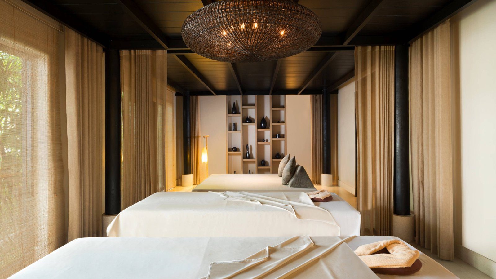 Spa 療程室裡三張白色按摩床，深色木樑天花板垂著藤編吊燈，四周是米色布簾
