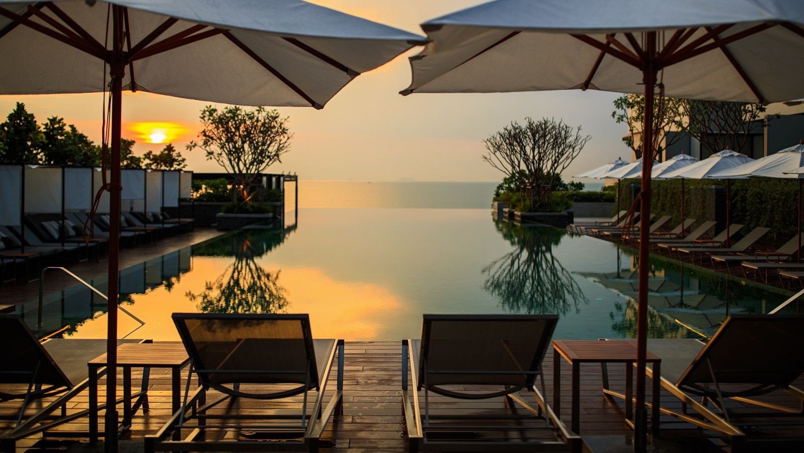 黃昏時分的泳池邊排著躺椅與遮陽傘，遠方是海面與夕陽