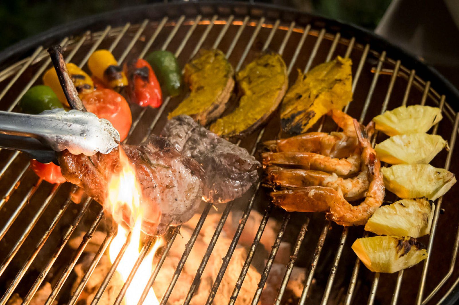 炭火烤架上正在燒烤牛排、鮮蝦、甜椒與鳳梨，夾子夾著一塊肉