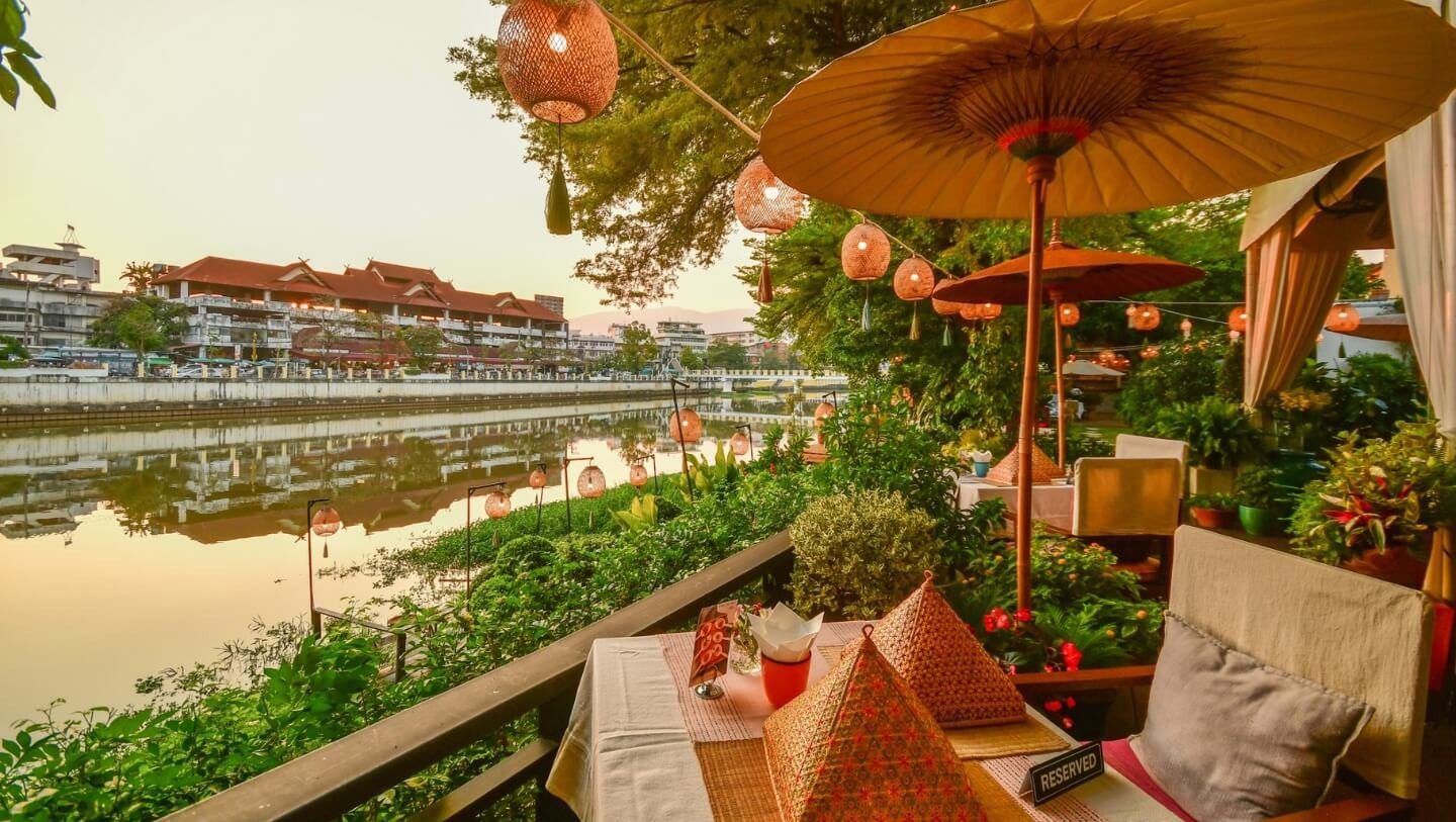 河畔露台掛著油紙傘與燈籠，擺著餐桌與坐墊，前方是平靜的河面