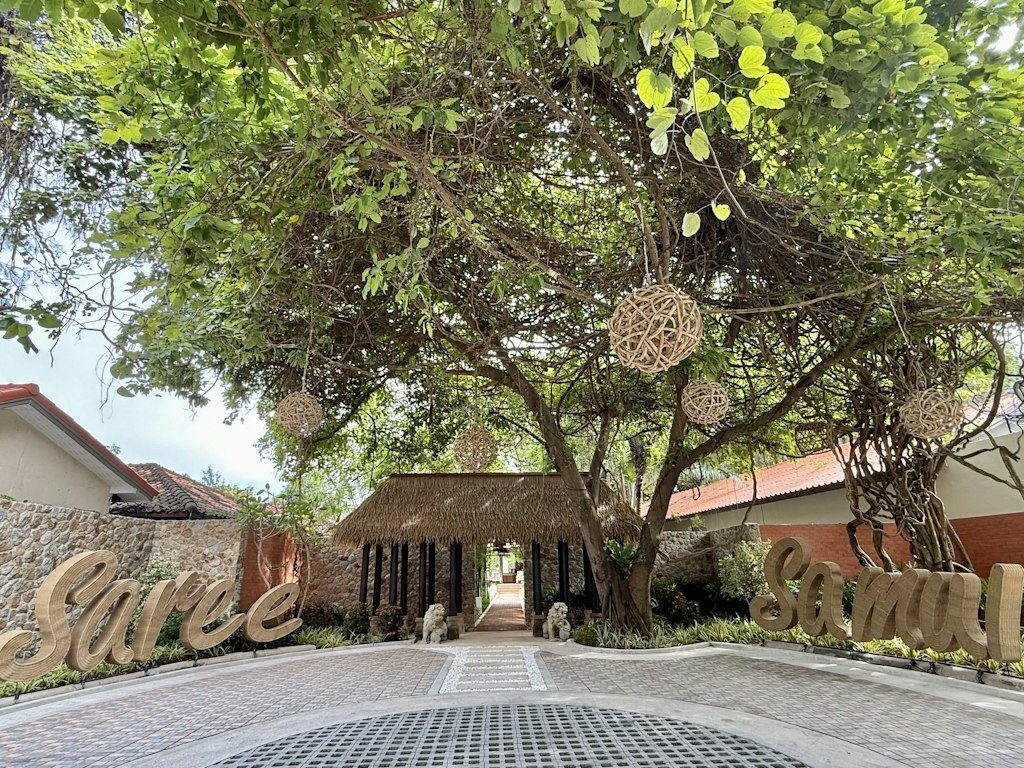 度假村入口的大樹下掛著藤球燈，地上是圓形鋪石車道，兩側有 Saree Samui 字樣的矮牆
