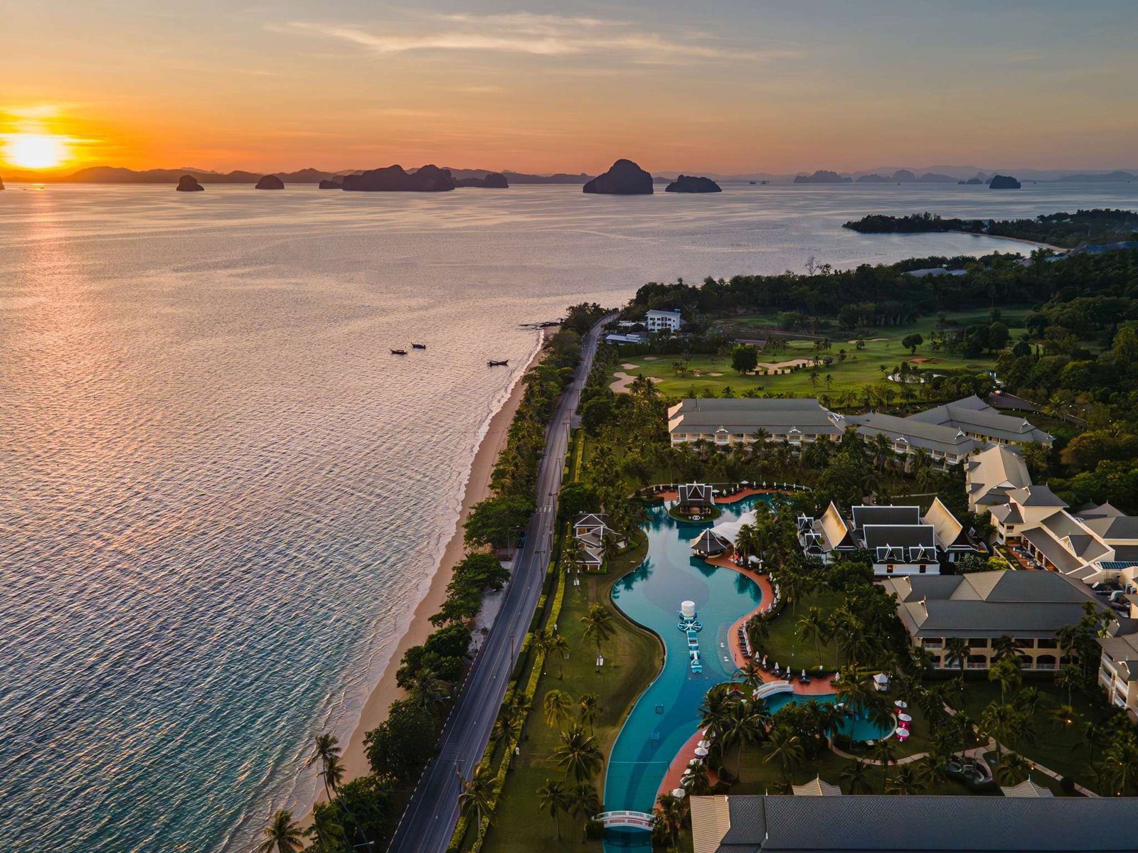 夕陽下的海岸線空拍，度假村與綠地沿著海灘延伸，海上有石灰岩島