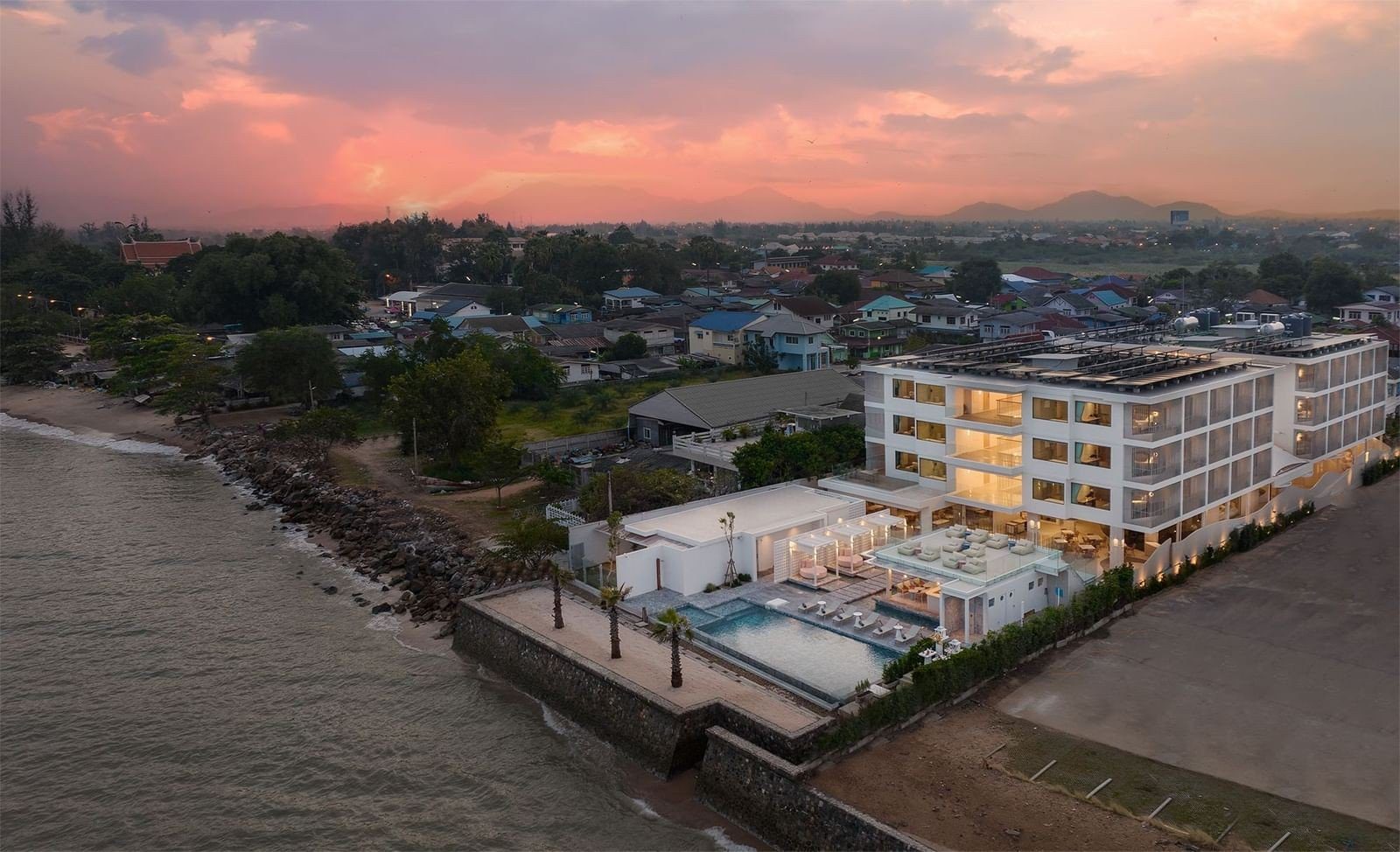 傍晚空拍海岸，白色飯店建築與泳池緊鄰海邊，後方是住宅區