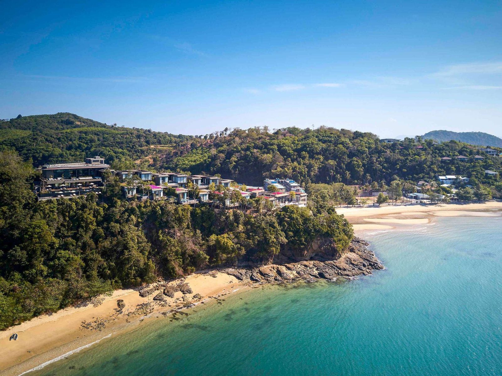 空拍樹林覆蓋的海岬，山坡上散布別墅建築，下方是一彎白沙灘與清澈淺海