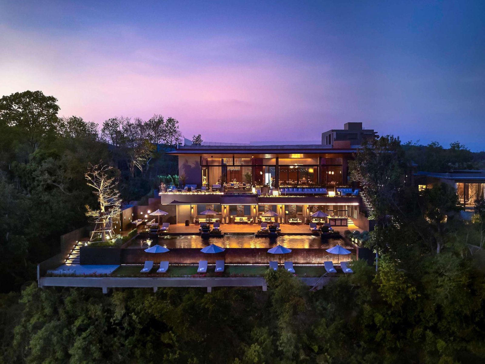 暮色中的山頂建築亮起燈光，多層玻璃立面與平屋頂藏在樹林間，背景是紫色晚霞
