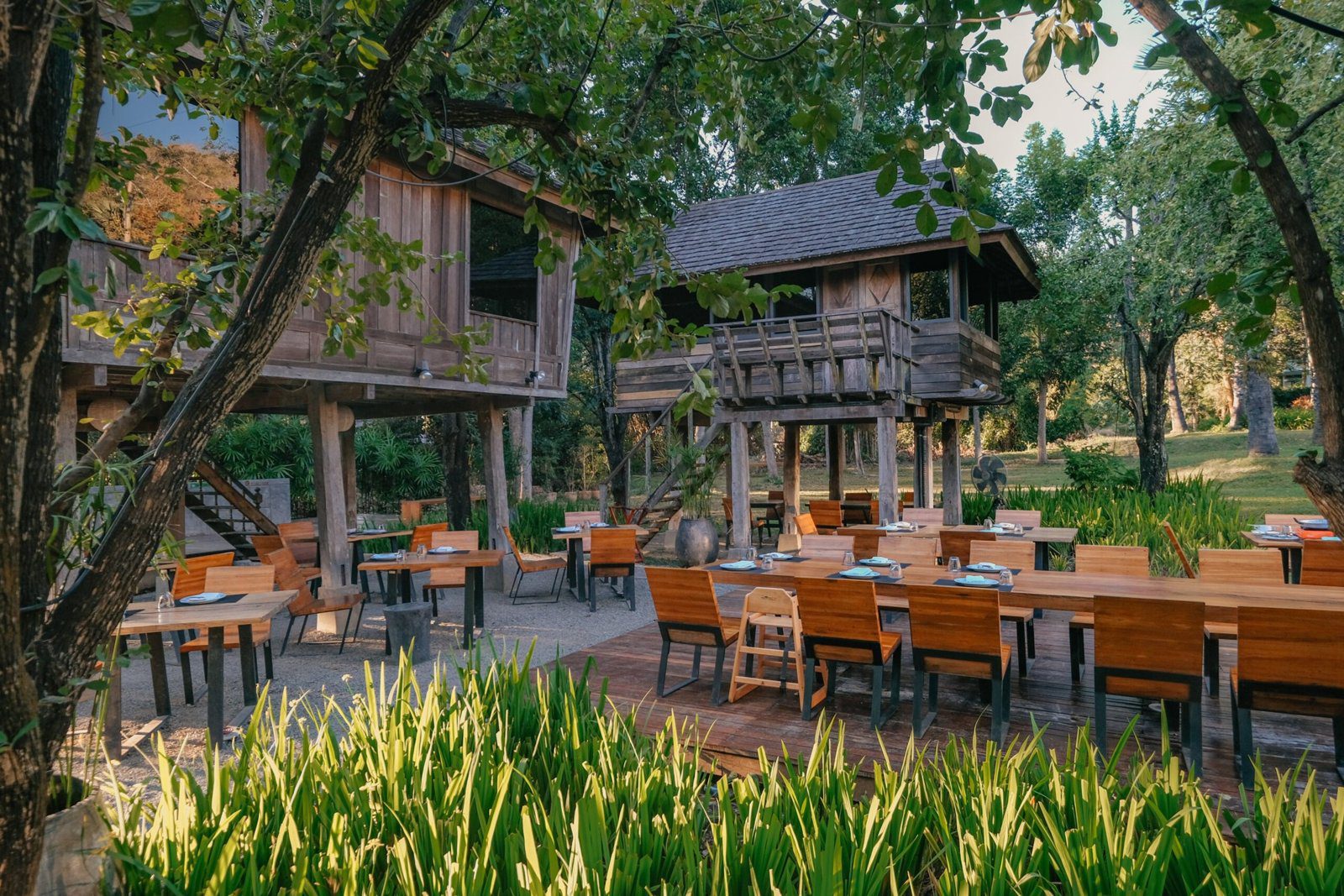 林間兩棟高腳木造米倉建築，前方木平台上排著餐桌椅，四周長滿綠色植物