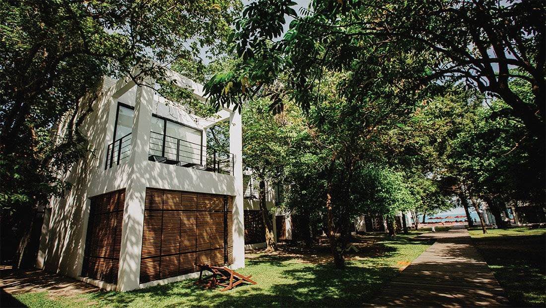 大樹林蔭下的兩層白色住宿建築，一樓是木百葉，旁邊的步道通往海邊