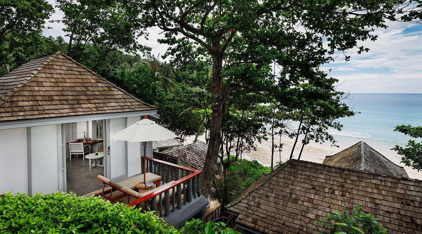 樹蔭下的白牆斜頂小屋與木頭露台，樹縫間看得到沙灘與海